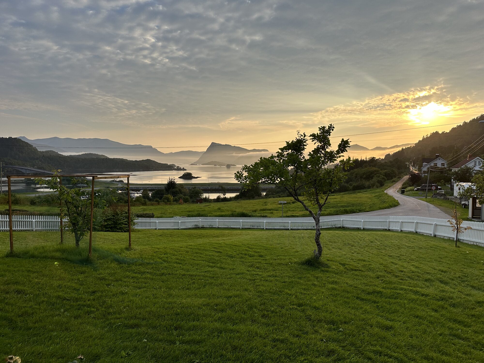 Kveldslys over ein hage med kvitt stakittgjerde og utsikt mot fjorden