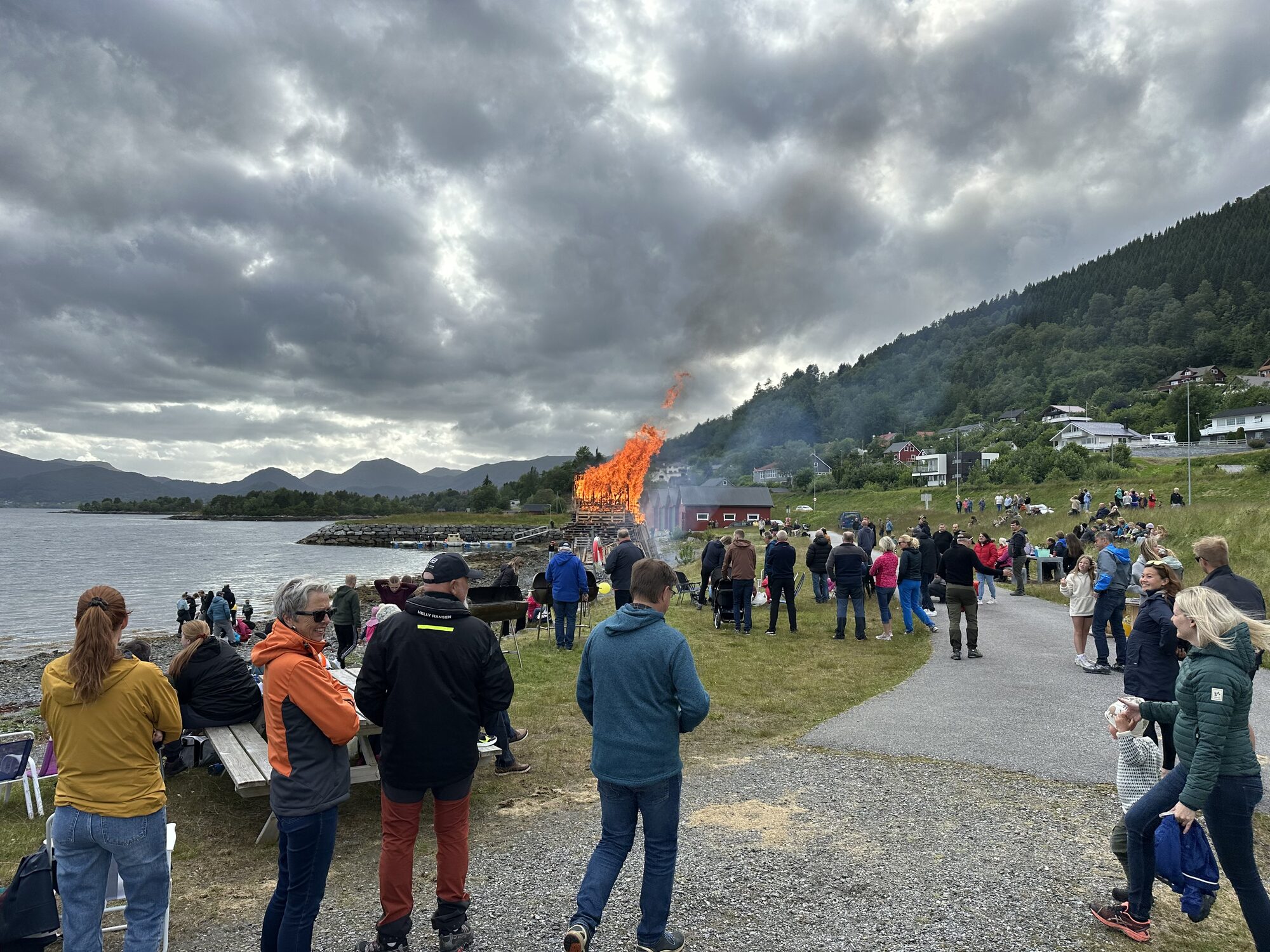 Stort sankthansbål brenn nede ved sjøen medan bygdefolk står samla rundt