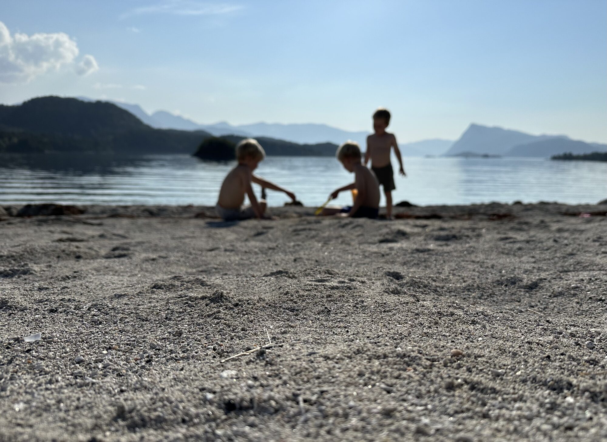Tre born leikar i sanden ved fjorden ein solrik sommardag